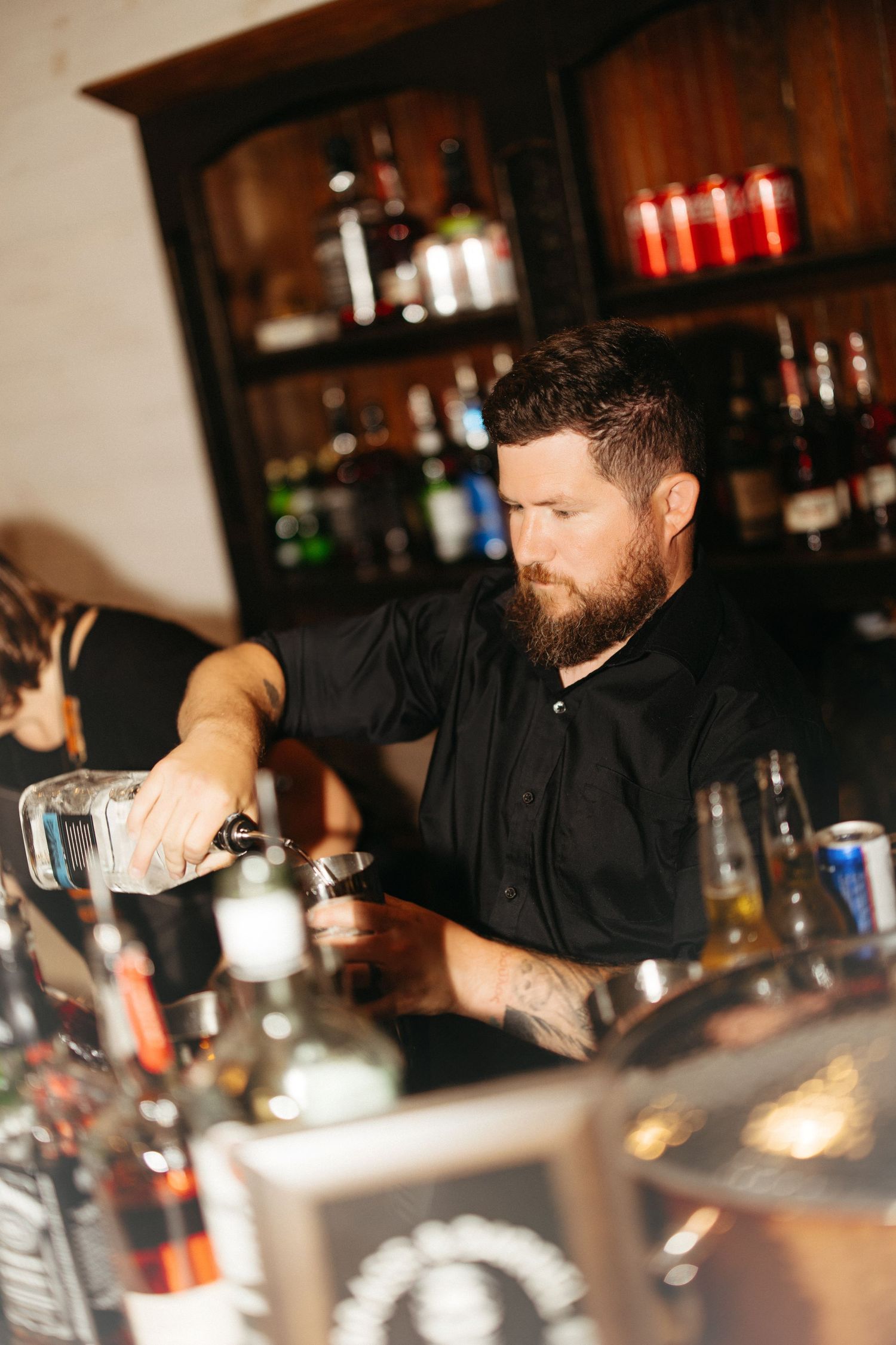 Professional bartender pouring drinks at wooden bar with bottles in background.