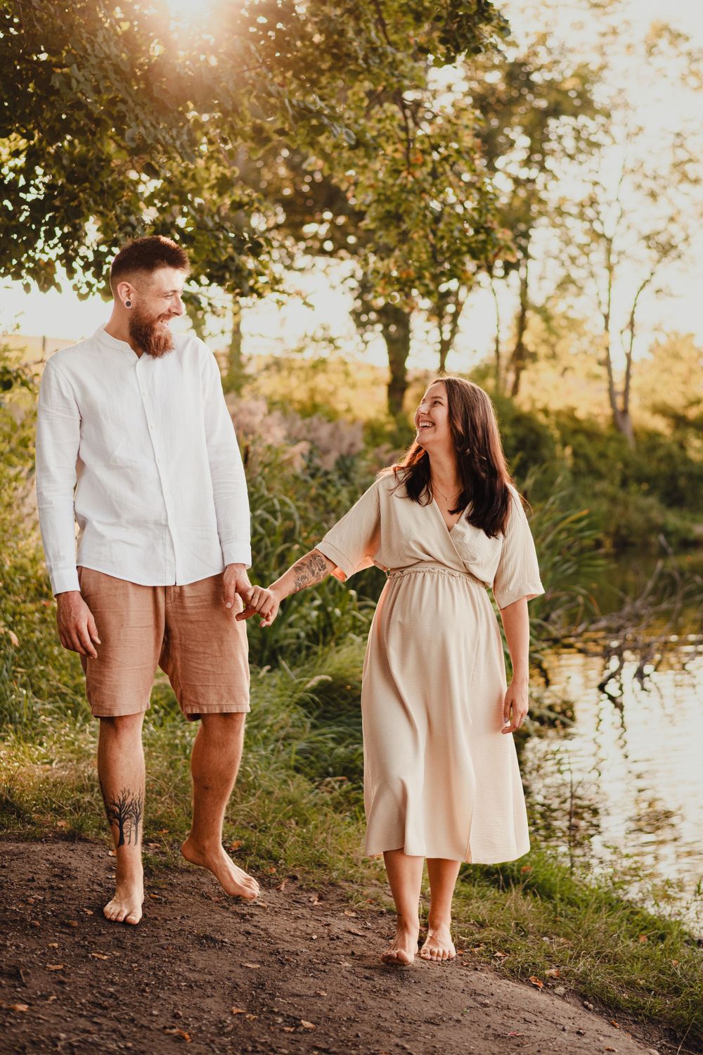 Couple in white and beige outfits walking barefoot along riverbank at sunset.