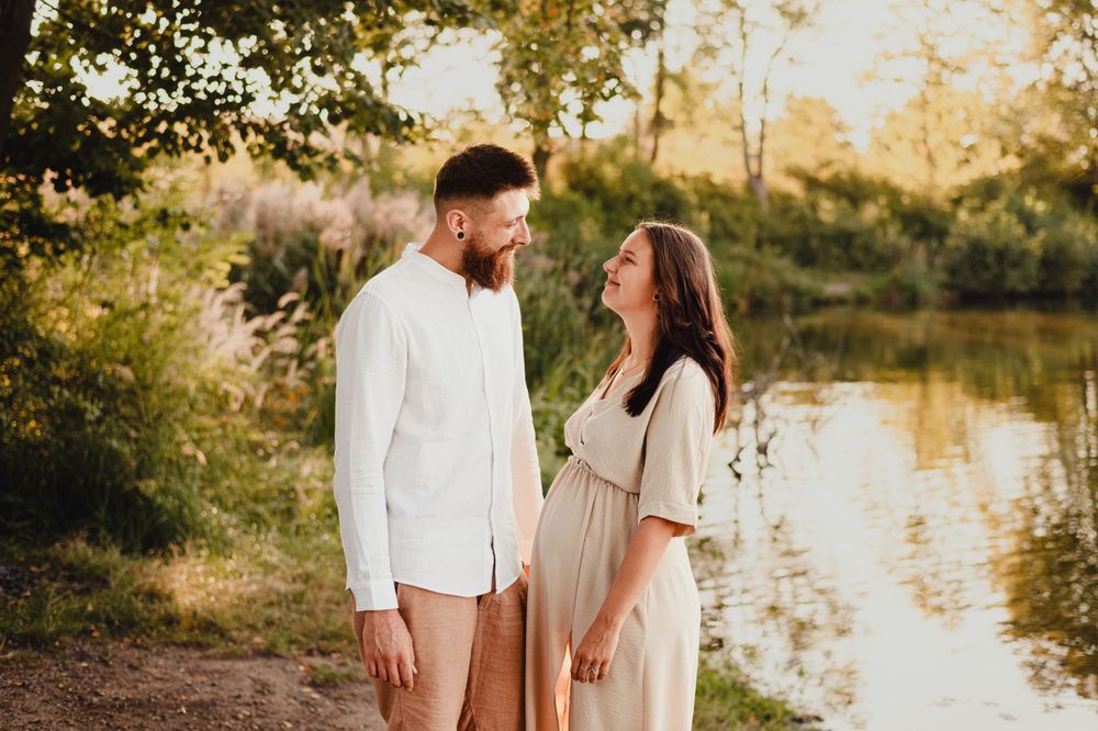 Romantic couple in light colored clothing embrace by river during golden hour.