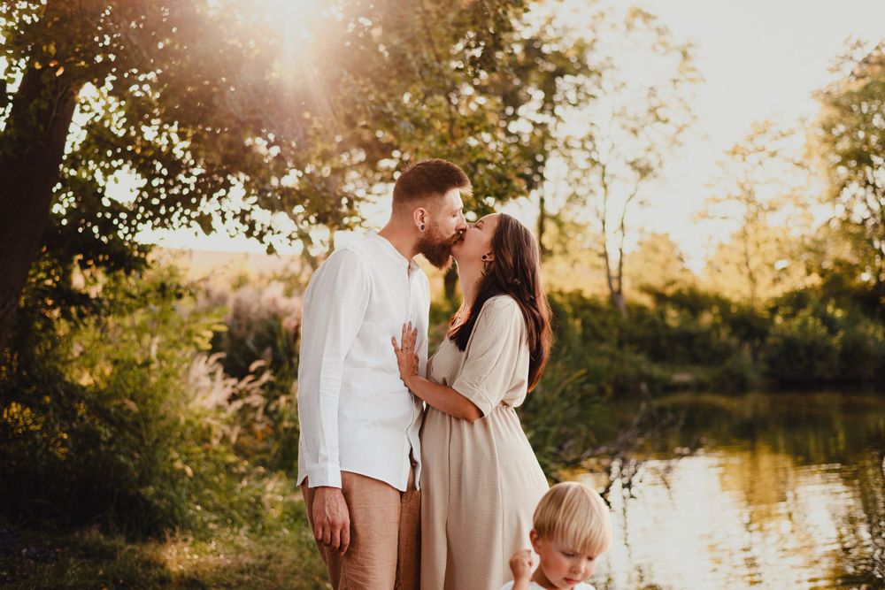Family of three in coordinating neutral outfits pose together by riverside at sunset.