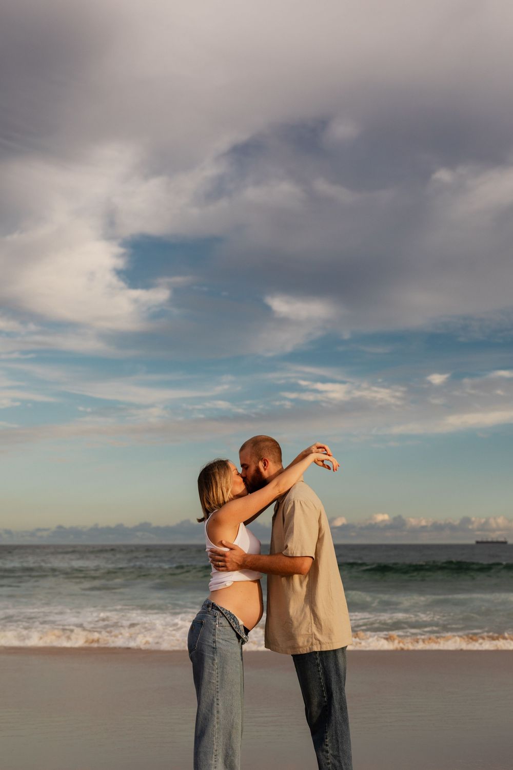 A couple embraces on a sandy beach with rolling waves and cloudy skies in the background.