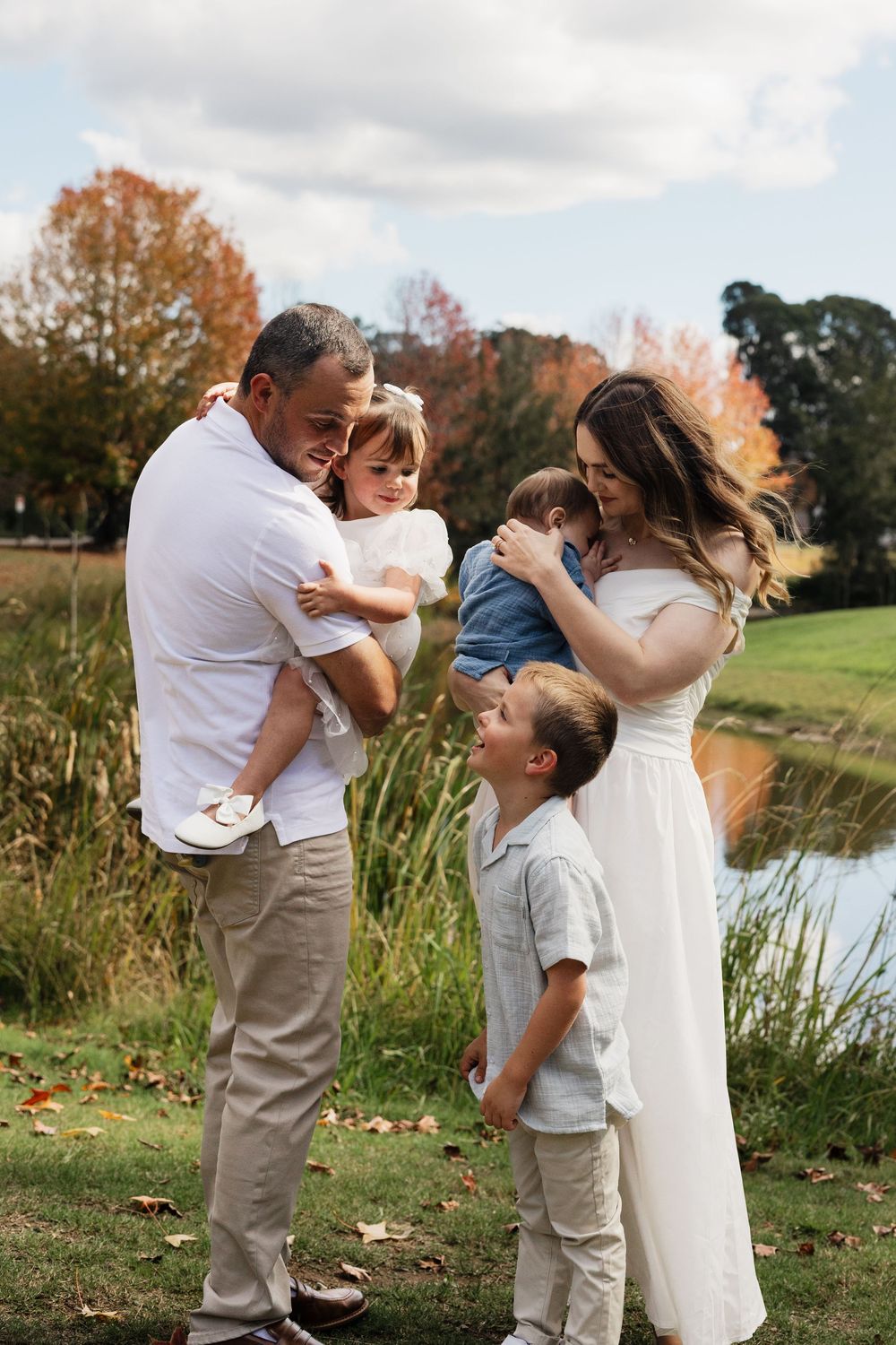 Family of five enjoying a fall day outdoors near a pond, parents holding children in autumn setting.