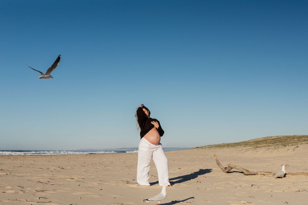Pregnant woman in white pants dancing on a sandy beach with a seagull flying overhead under a clear blue sky.
