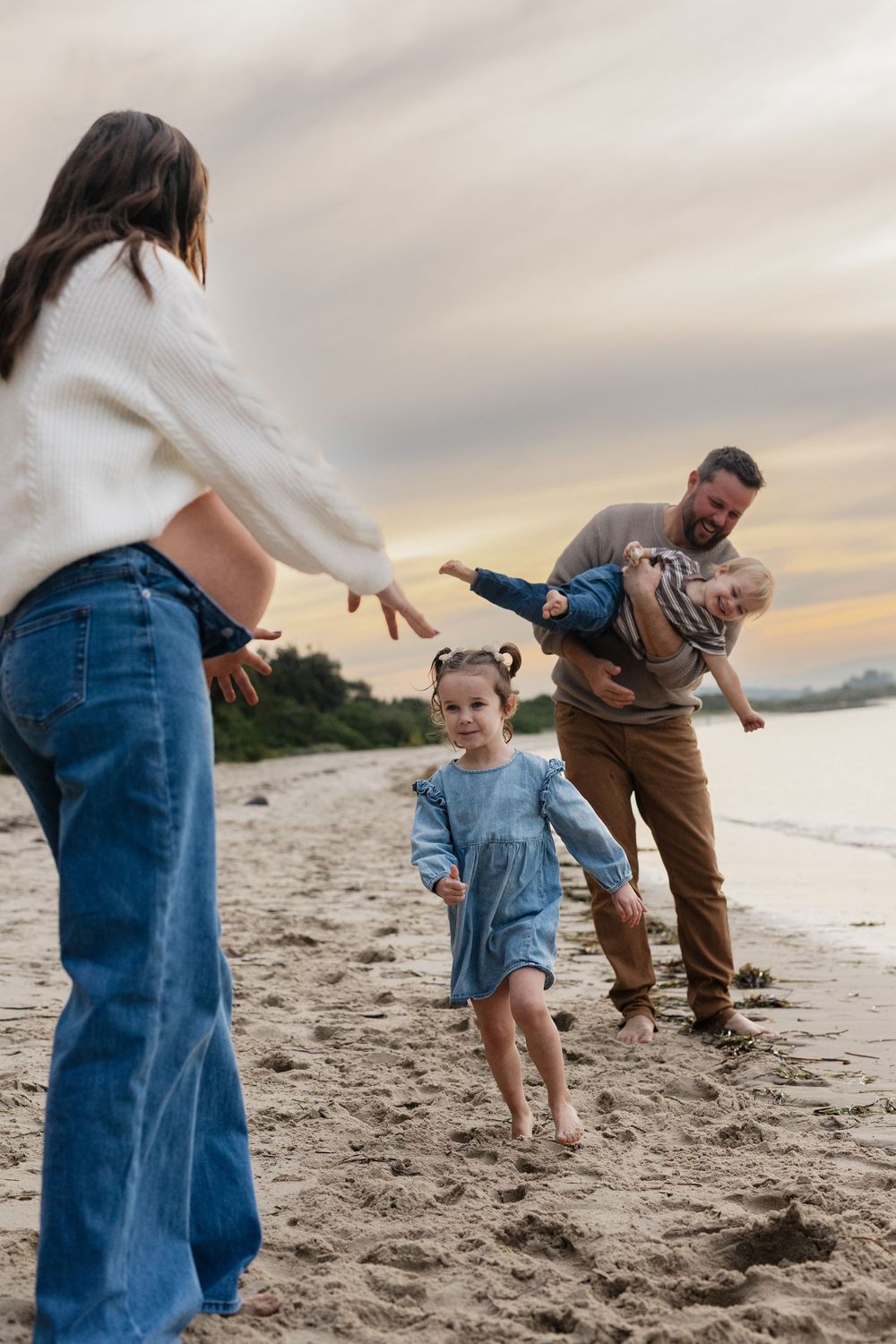 Family playing on sandy beach at sunset, father holding child while daughter runs to mother in white sweater.
