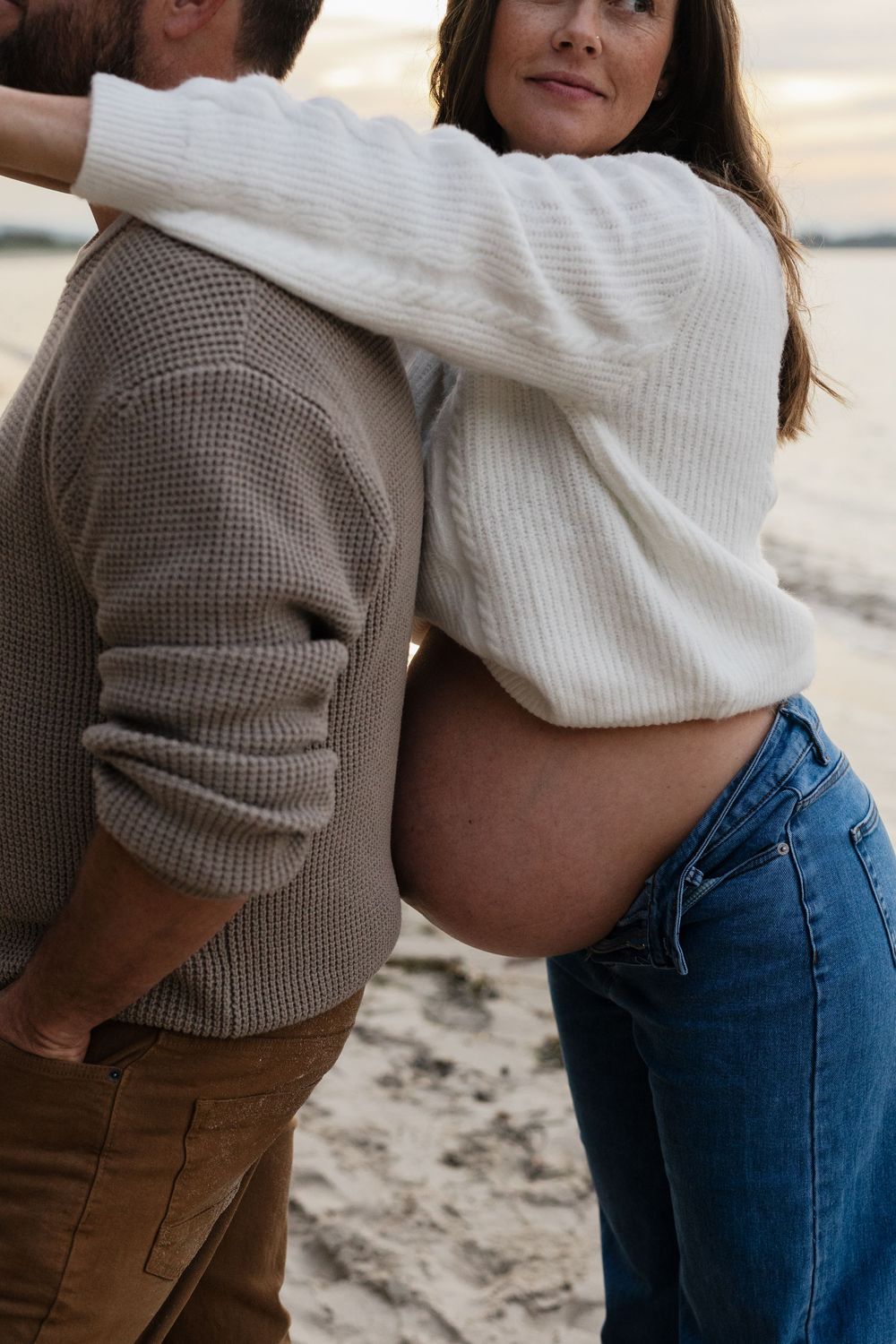 Pregnant woman in white sweater and jeans embraces partner on sandy beach at sunset.