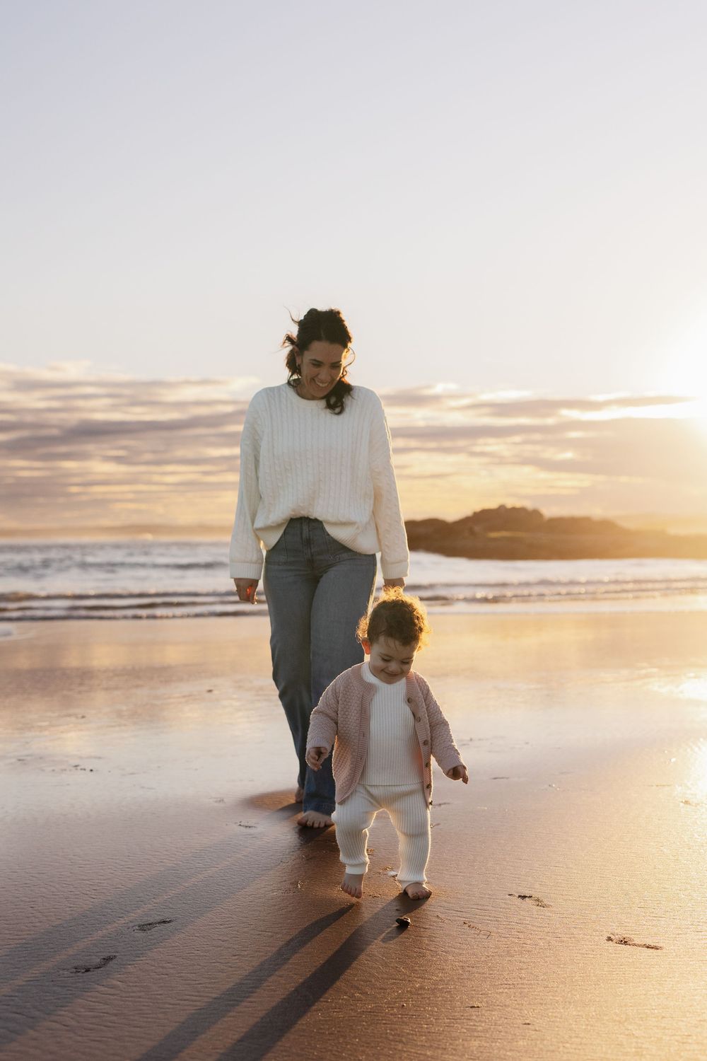 A mother and toddler walk barefoot on a sandy beach at sunset, with golden light reflecting on the wet sand.