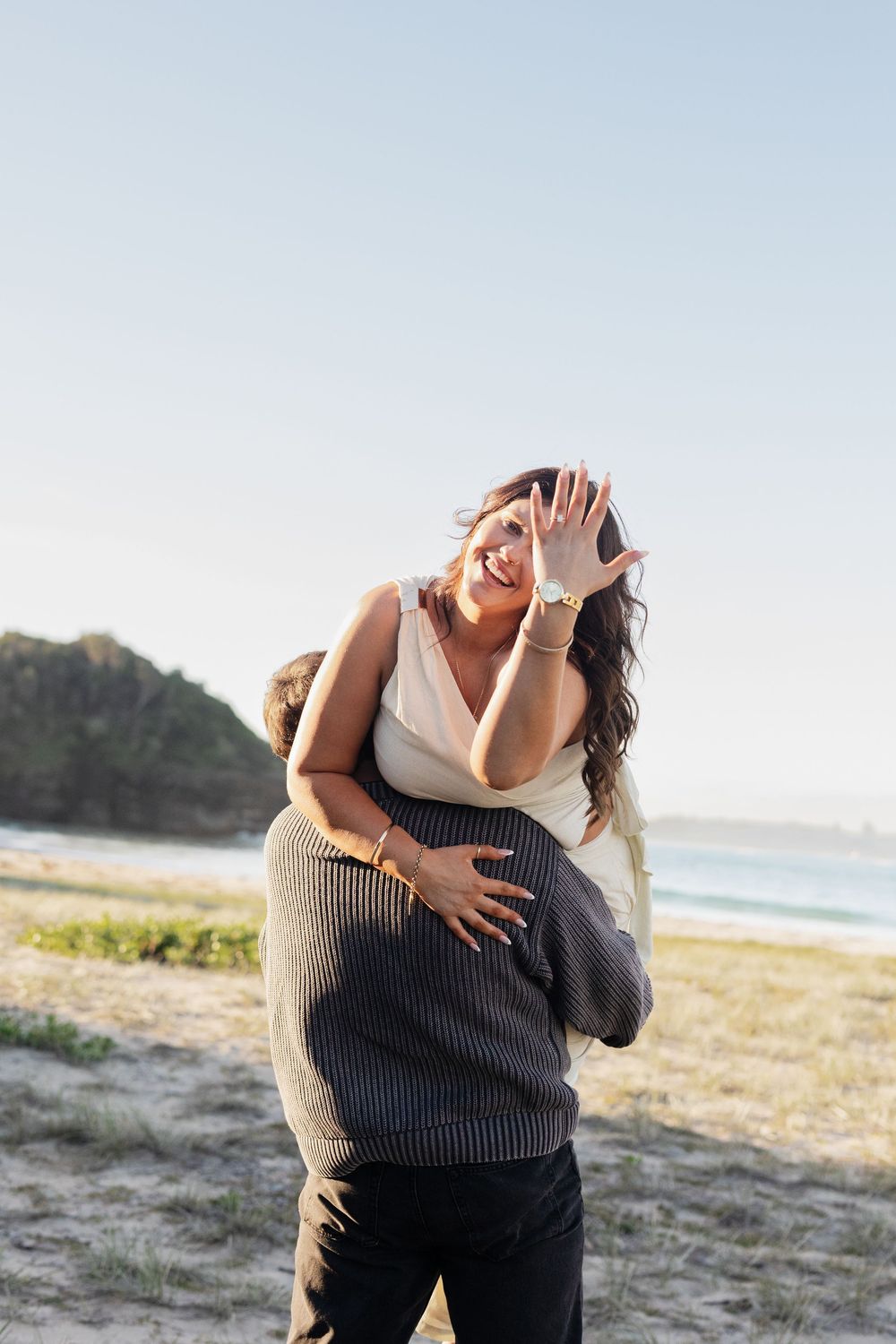 A joyful couple embracing on a beach, woman laughing and waving at camera, ocean and cliffs in background.
