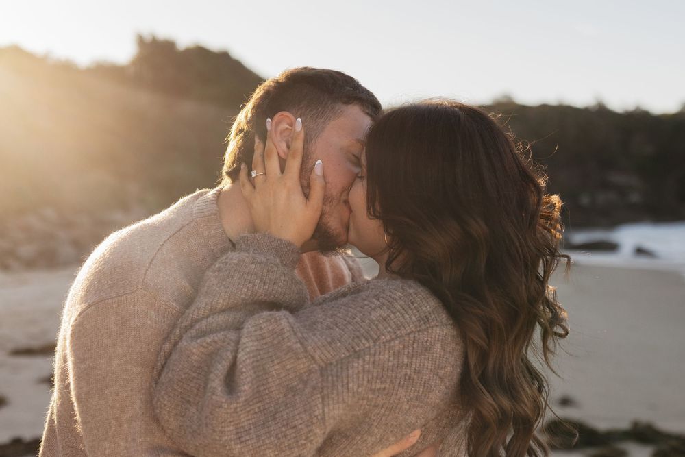 Couple sharing a romantic kiss on a beach at golden hour, both wearing cozy knit sweaters.