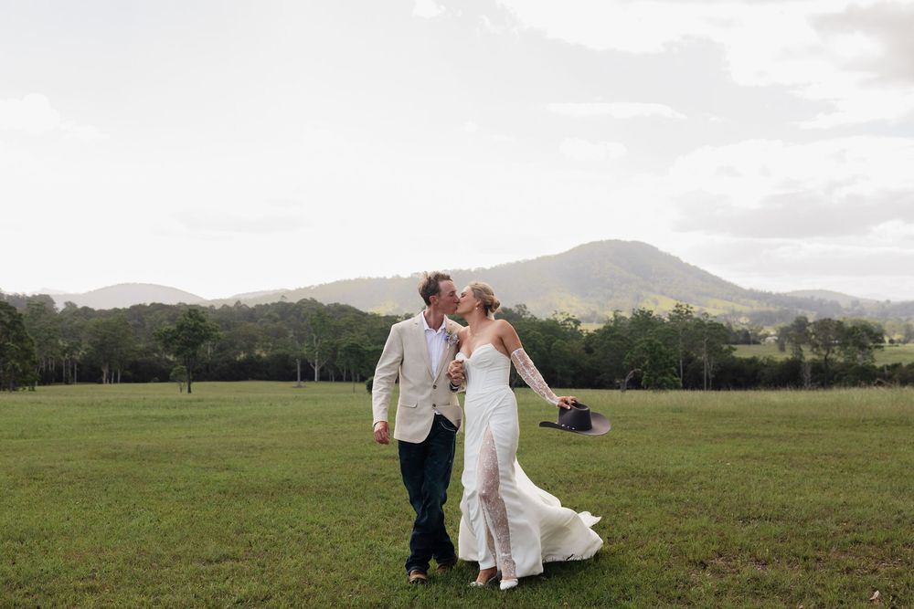A couple shares a romantic kiss in an open green field with mountains in the background.