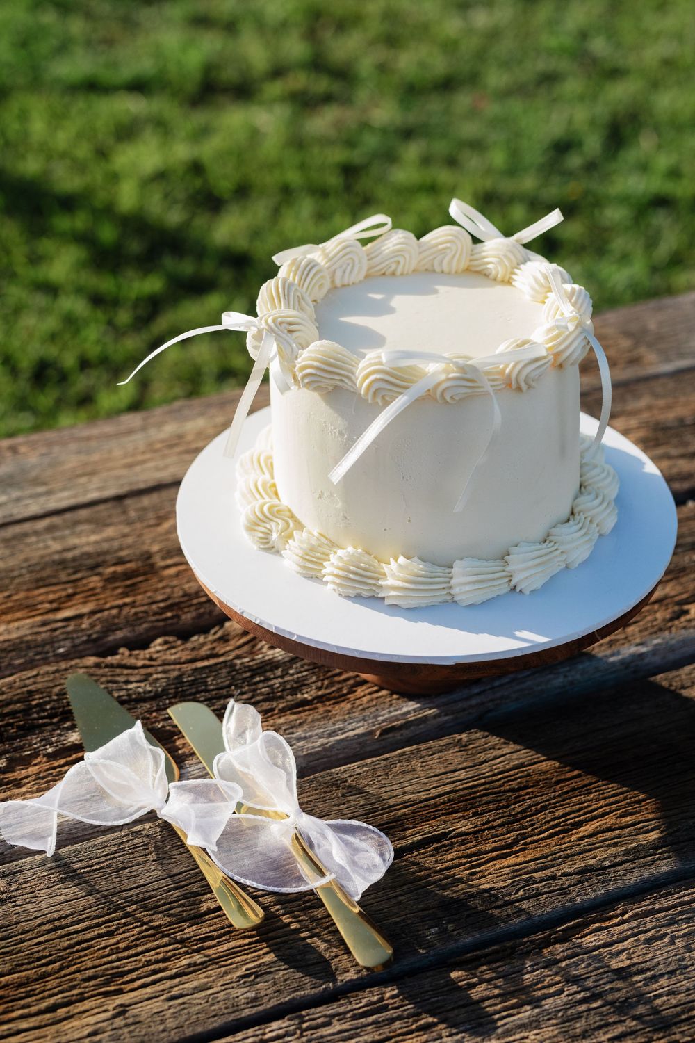 Elegant white wedding cake with delicate piping and ribbon decorations sits on rustic wooden table with serving utensils.