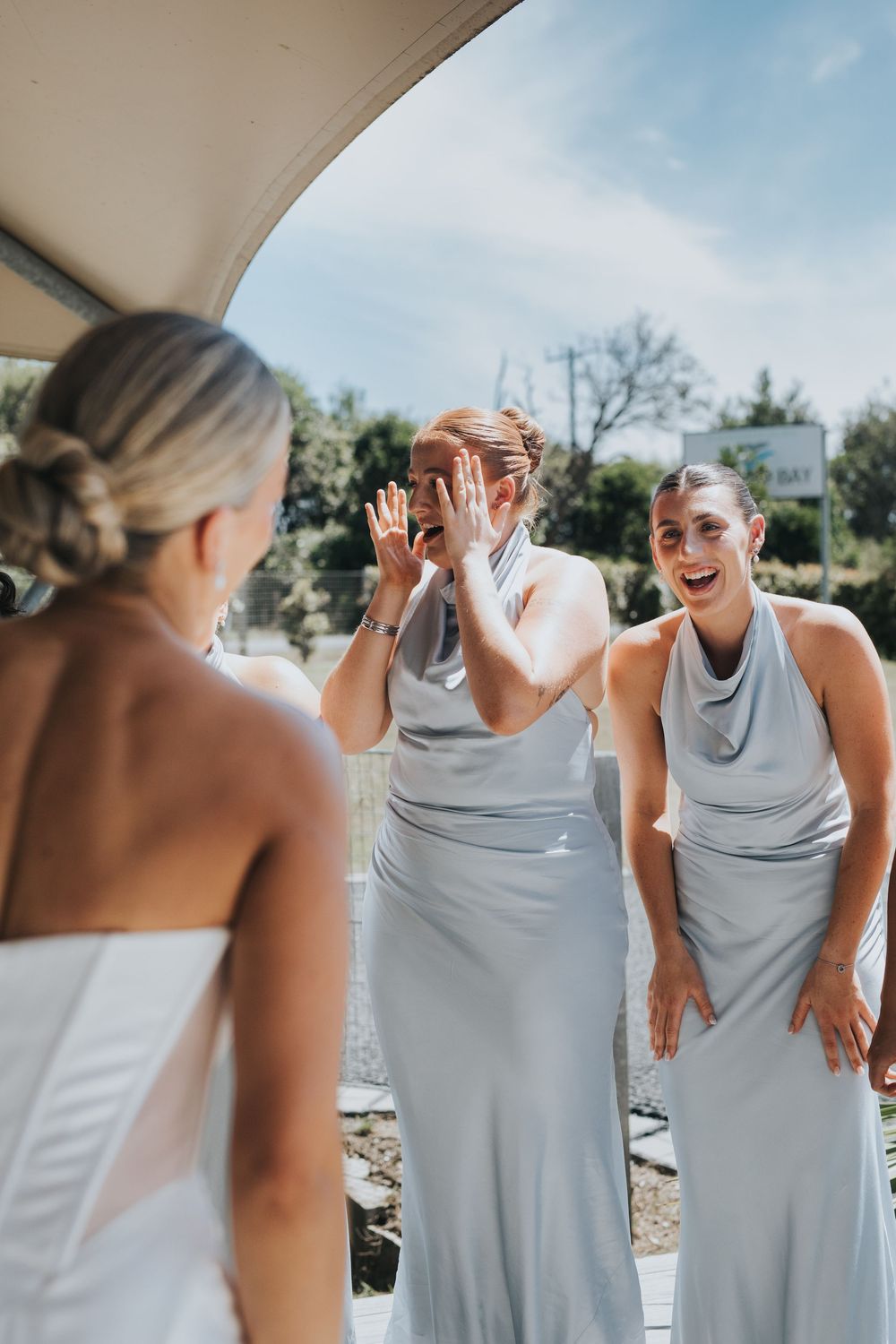 A group of bridesmaids in light gray dresses share candid laughing moments during a wedding photoshoot outdoors.