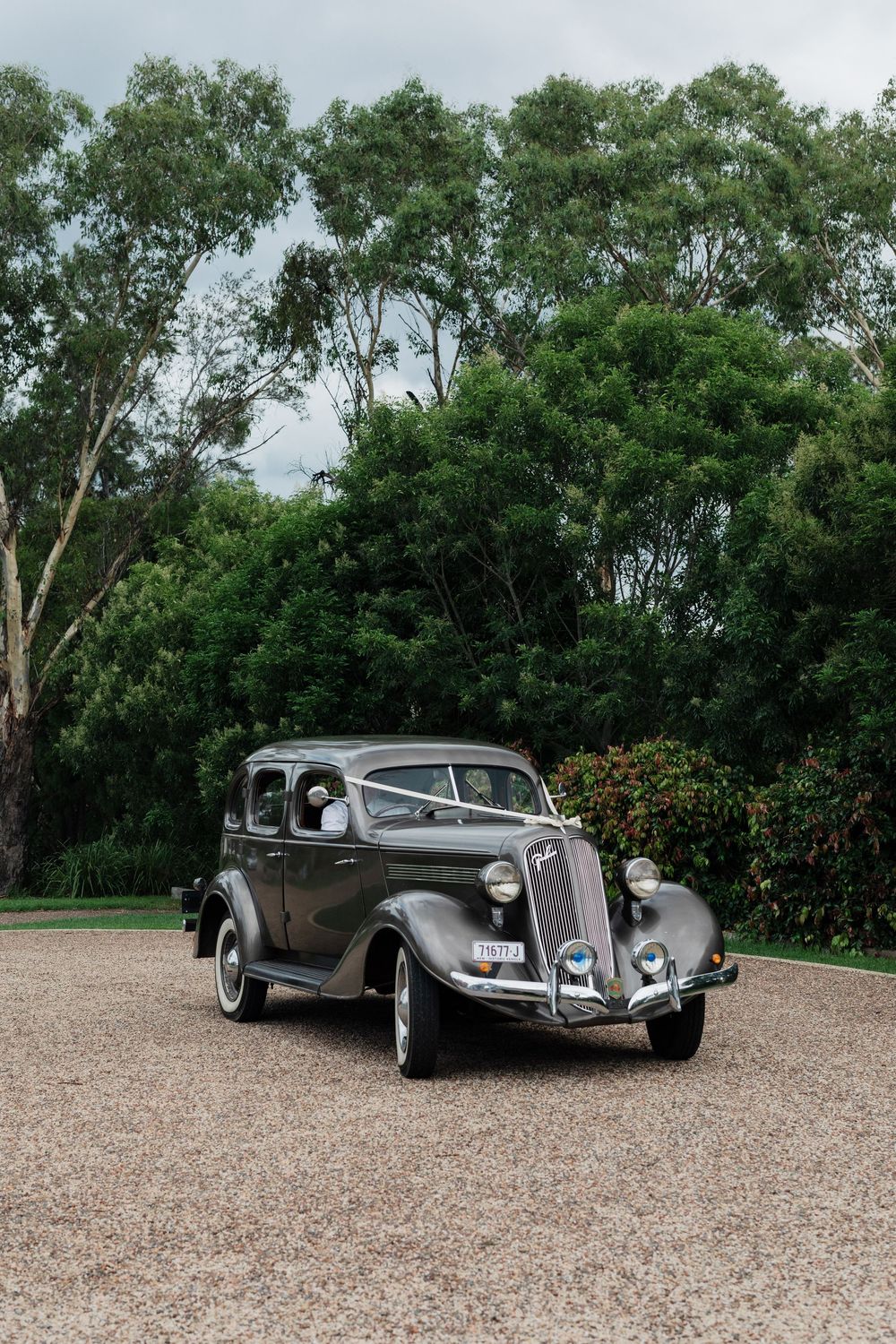 Three classic luxury cars from the 1930s displayed on a gravel surface with lush green trees in the background.