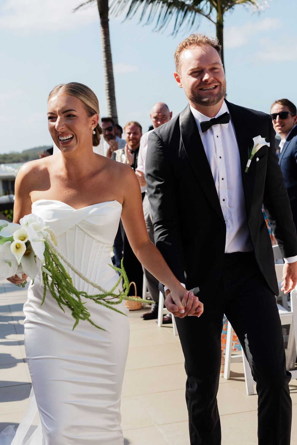Newlyweds share a joyful moment walking together at tropical beach wedding with palm trees in background.