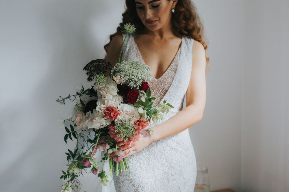 Bride in lace wedding dress holds elegant bouquet of burgundy and pink flowers against light grey backdrop.