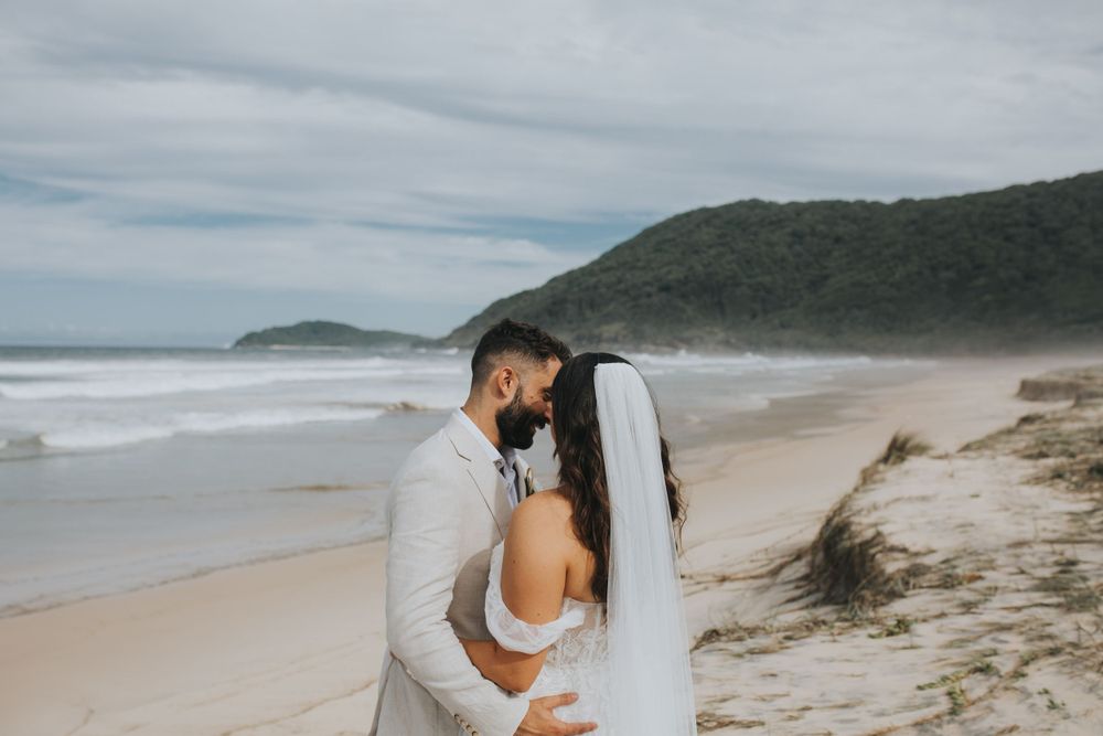 Couple in wedding attire embracing on a scenic beach with waves in background.
