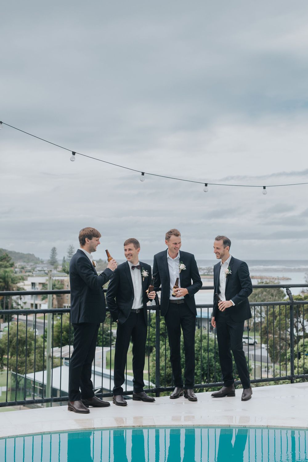 A group in formal attire sits along the edge of a rooftop pool with string lights and city views in the background.