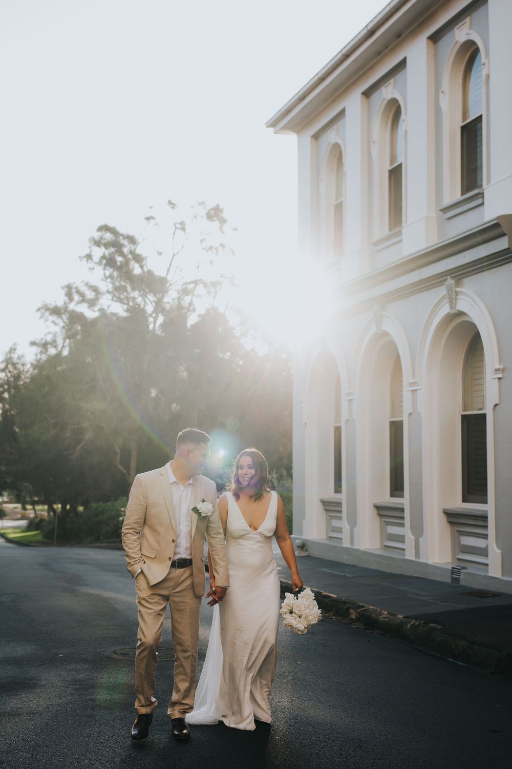 Bride and groom walk hand-in-hand outside a white colonial building, bathed in golden sunlight with a stunning lens flare.