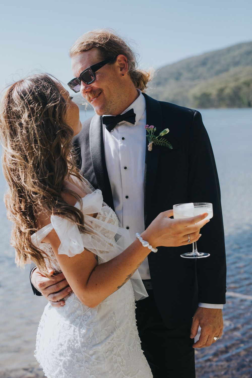 A couple in wedding attire share a romantic moment by a lakeside, holding champagne glasses and smiling at each other.