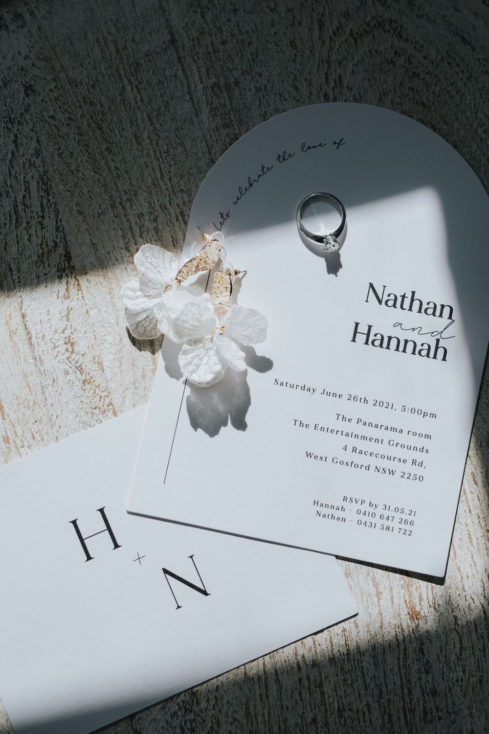 Wedding invitation with white flowers and engagement ring photographed in dramatic lighting on a textured surface.