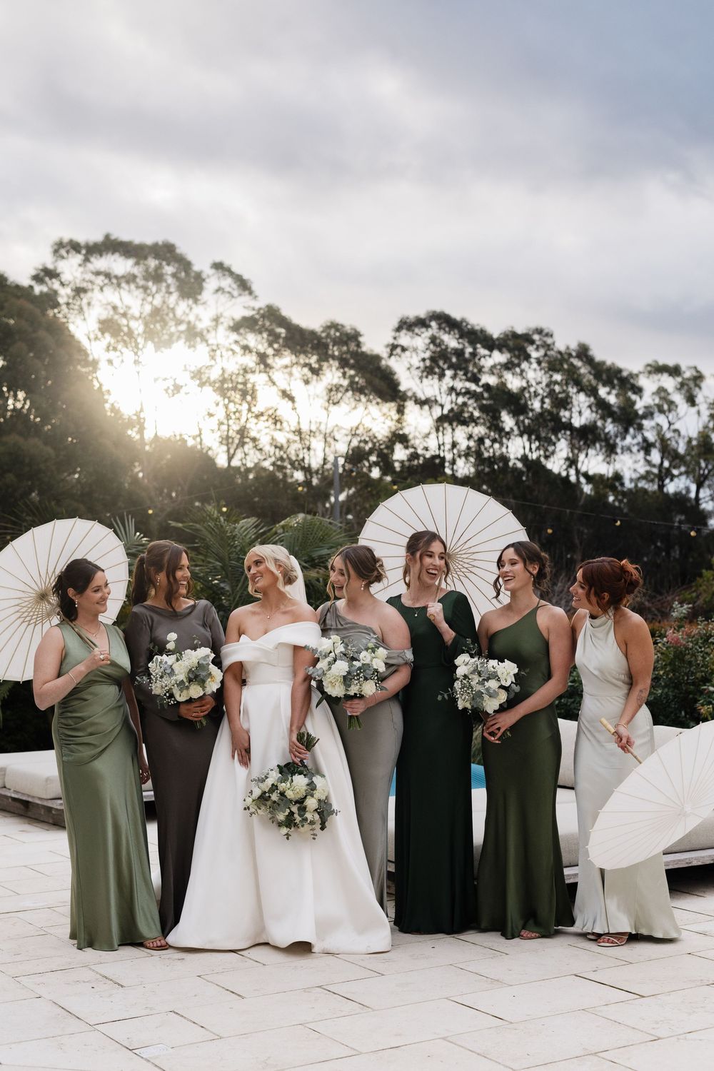 Bride in white off-shoulder gown with bridesmaids in green dresses holding white parasols and bouquets outdoors.