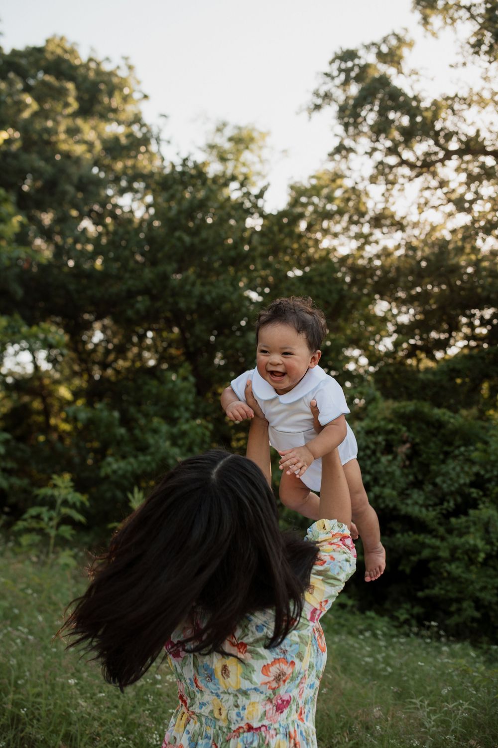 A parent lifts a laughing baby wearing white into the air against a green outdoor backdrop at sunset.