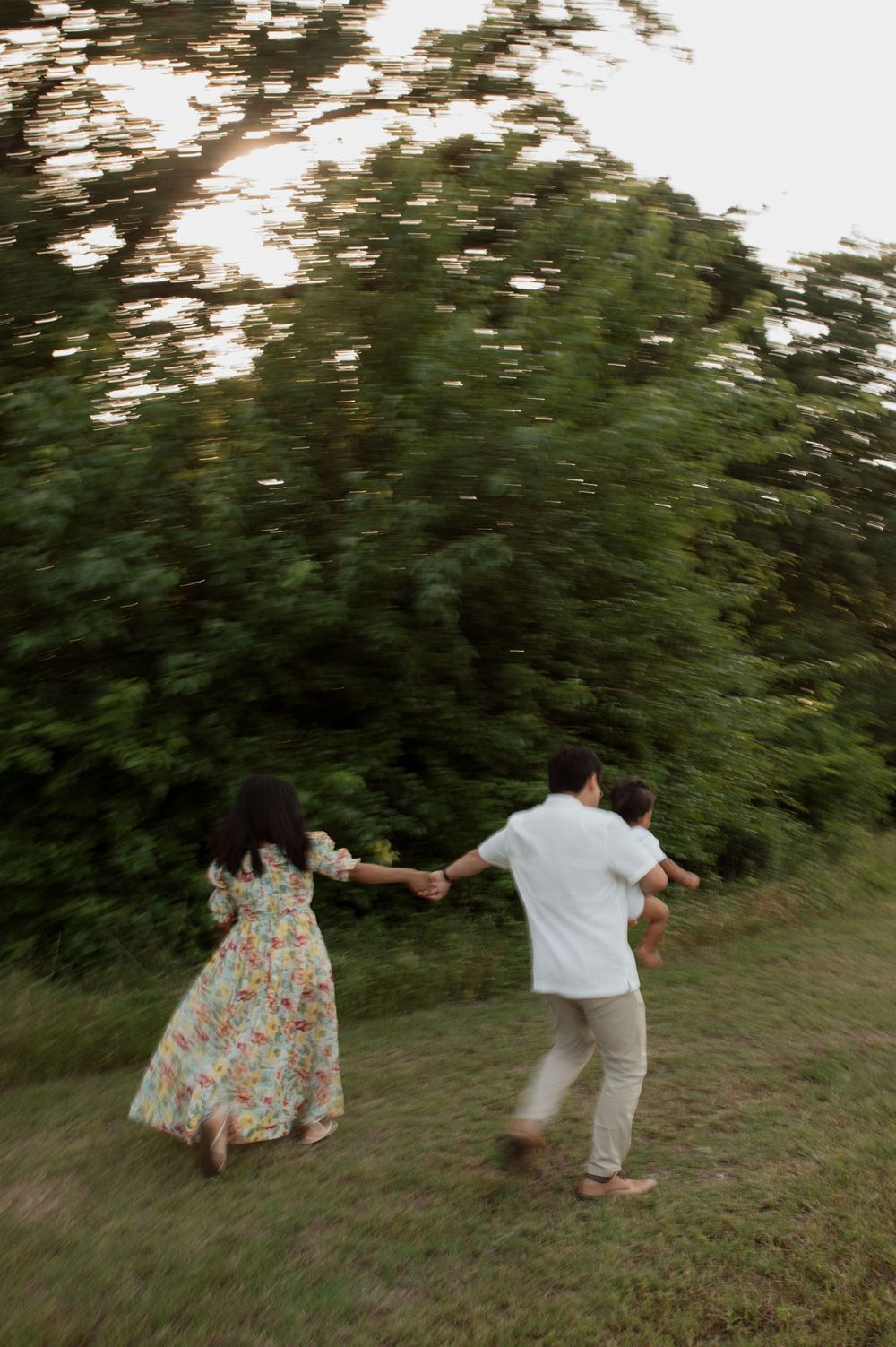 Two people hold hands while dancing and spinning together on a grassy hillside at sunset with trees in the background.