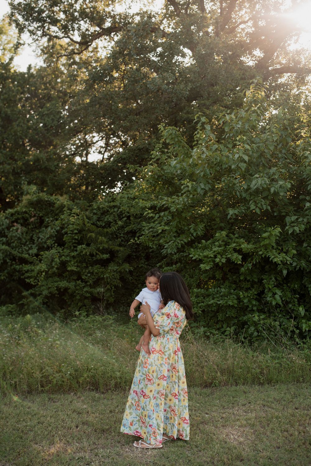 A person in a floral dress holding a small child while standing in a sunlit garden setting at dusk.