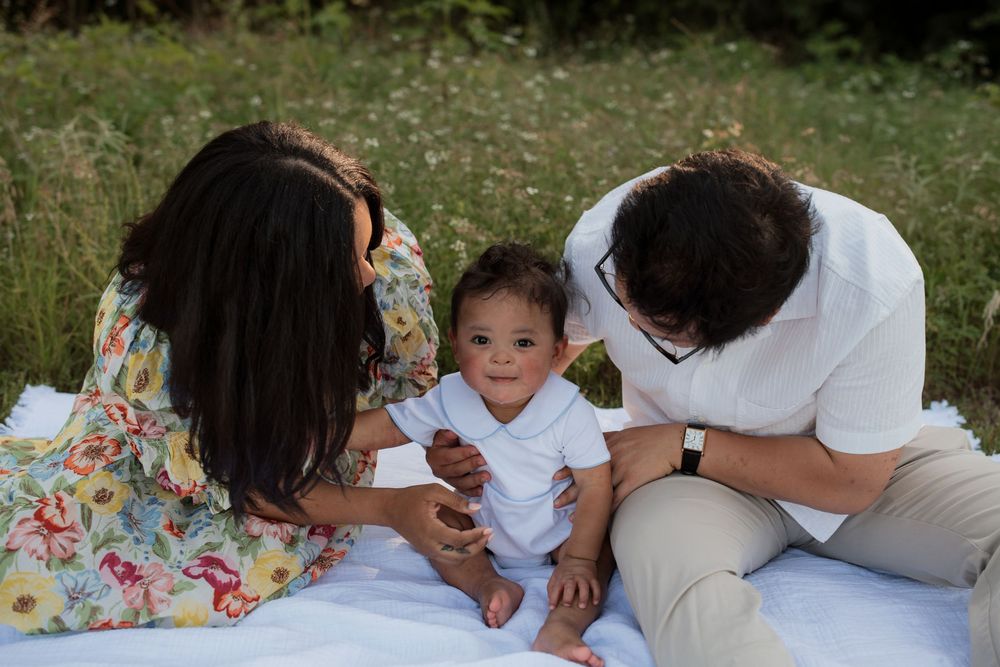 A family shares a tender moment together while relaxing on a white picnic blanket in a grassy field.