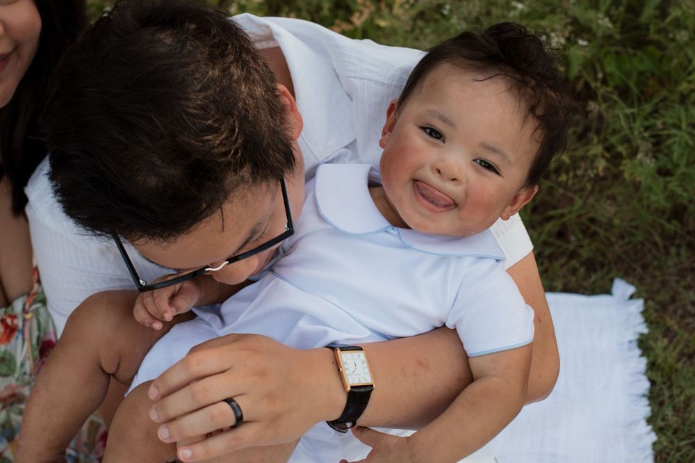 A happy child in a white outfit smiles while being held outdoors on a sunny day.