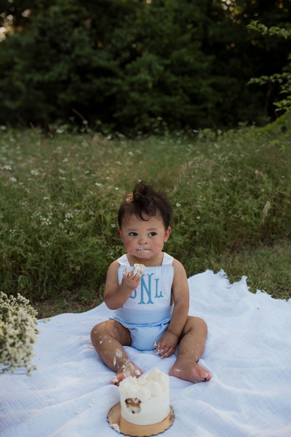 Small child in light blue romper sits on white blanket with smash cake during outdoor first birthday photoshoot.