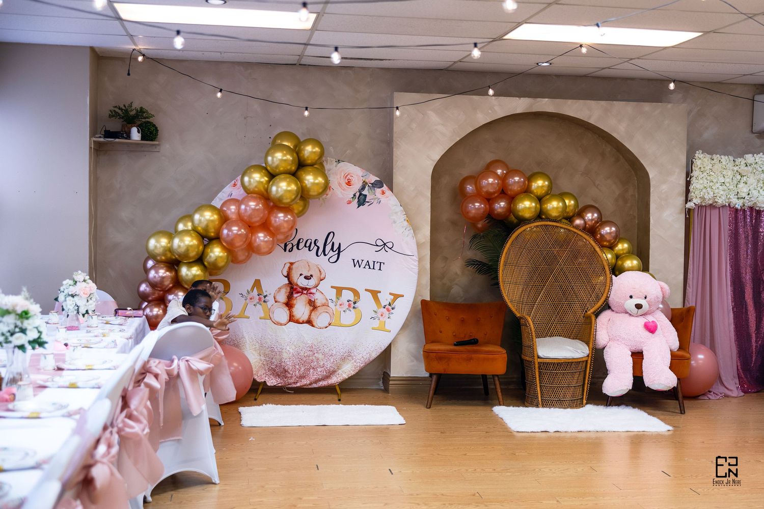 Baby shower decoration setup with balloon arch, throne chair and pink teddy bear.
