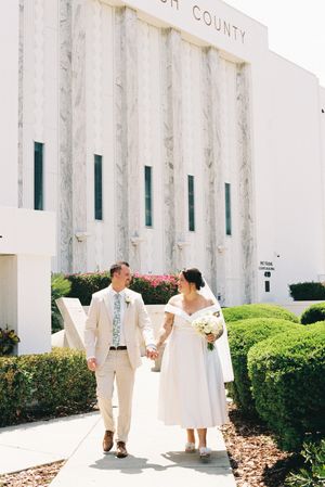 A couple in white wedding attire holding hands while standing outside a white building with green landscaping.