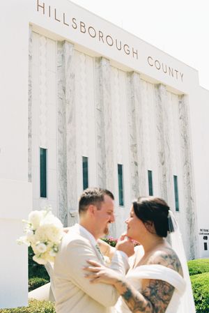 A wedding couple embraces in front of the Hillsborough County building with white flowers and classic architecture.