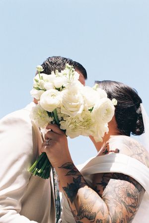 A couple shares a kiss behind a bouquet of white roses against a light blue sky background.