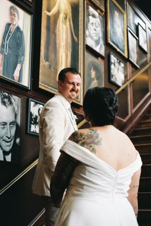 A couple in wedding attire stands on wooden stairs near a wall decorated with vintage black and white photographs.
