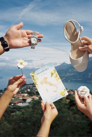 Hands holding wedding details including a passport program, ring box, white sandal, and watch with Italian lake views.