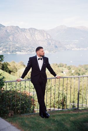Dapper groom in black tuxedo leaning on railing with breathtaking lake and mountain scenery behind him.