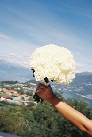 Bride holding a bouquet of white roses against a blue sky and lake landscape.