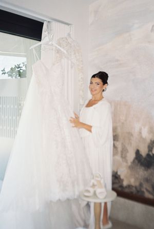 Smiling bride in white robe admires her elegant lace wedding gown hanging by a window with bridal heels nearby.