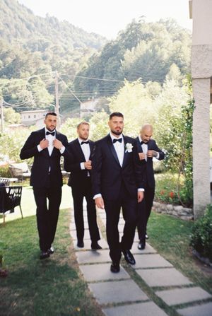 Groom and groomsmen in black tuxedos walking confidently along a garden path at an outdoor wedding venue.
