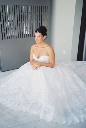 Bride in a voluminous strapless lace ball gown sitting elegantly indoors.
