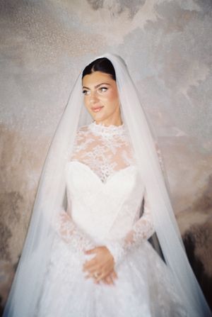 Stunning bride in elegant lace high-neck wedding gown with cathedral veil gazing upward in soft light.