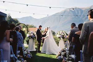 Outdoor wedding ceremony with bride and groom at floral arch, guests seated, string lights, and scenic mountain backdrop.