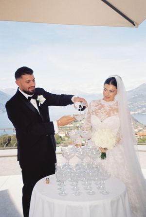 Bride and groom pouring champagne into a classic tower of glasses at an outdoor wedding with scenic lake views.