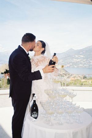 Newlyweds kissing while pouring champagne tower at their elegant lakeside wedding reception in Italy.
