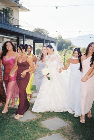 Joyful bride in lace gown walks with colorful bridesmaids in mismatched dresses at an outdoor wedding.