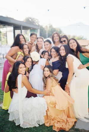 Bride surrounded by bridesmaids in colorful dresses, smiling together at an outdoor wedding reception.