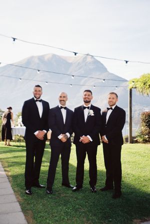 Four groomsmen in black tuxedos posing outdoors with mountain backdrop at elegant wedding venue.