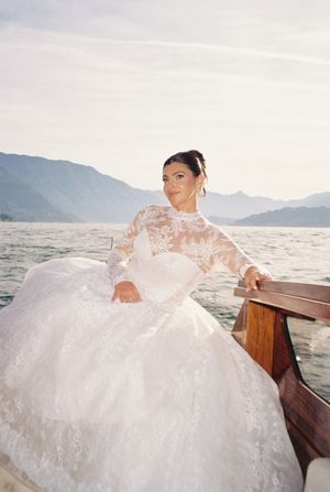 Bride in lace ball gown sits on wooden boat with alpine lake backdrop.