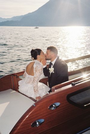 Bride and groom sharing a romantic kiss on a wooden boat with sunlit Lake Como and mountain backdrop.